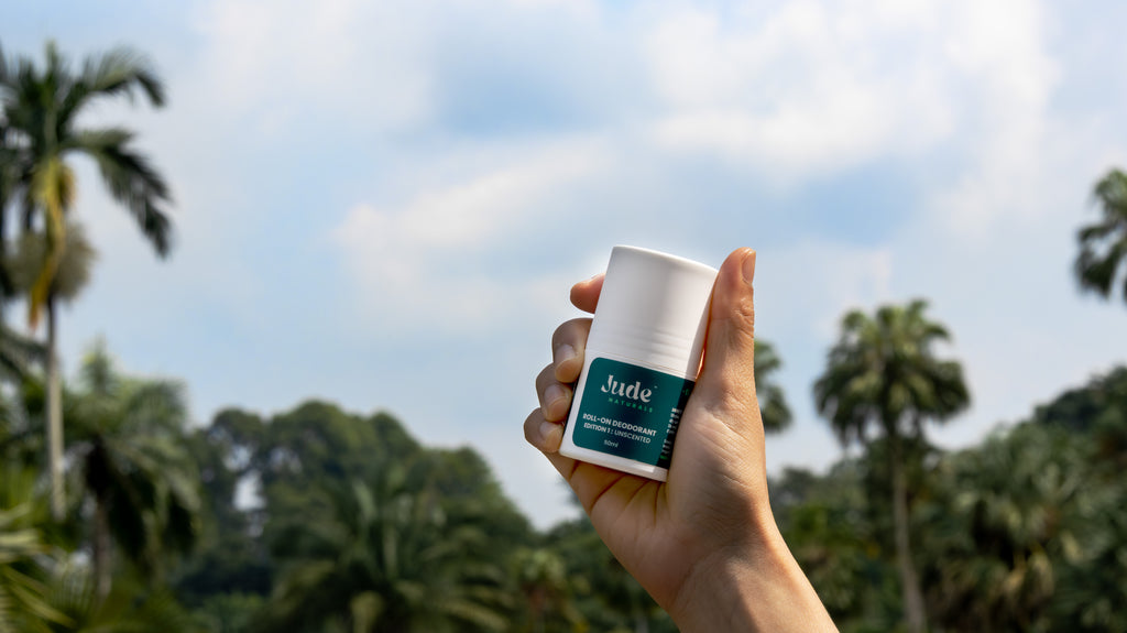 Human hand holding 50ml bottle of Jude Naturals Unscented natural roll-on deodorant. Live background of palm trees. 