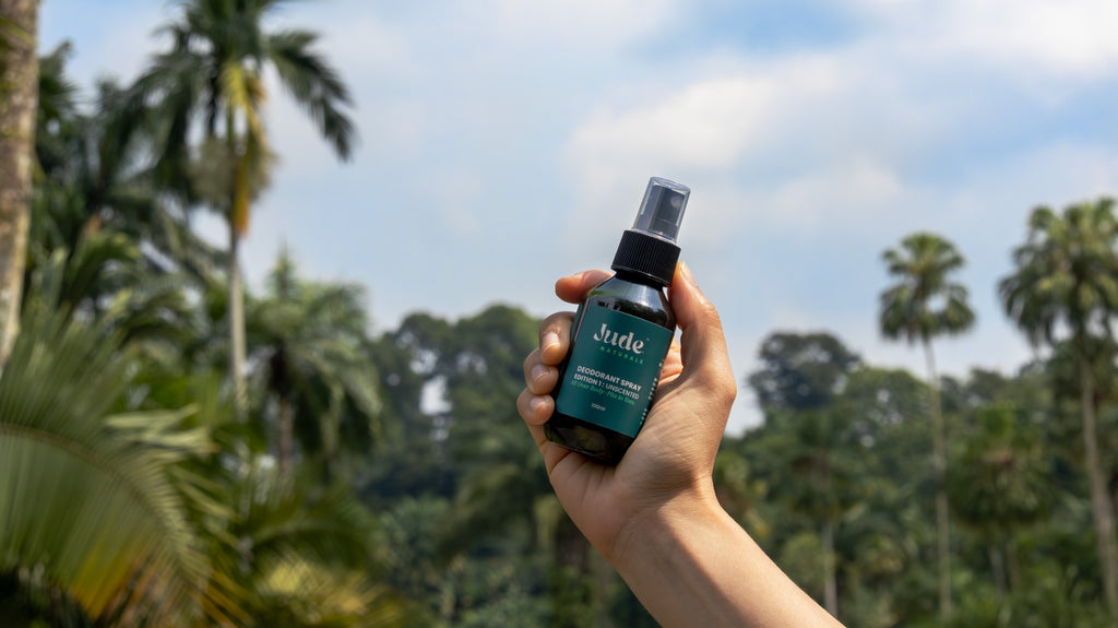 Human hand holding 100ml Jude Naturals spray deodorant with mineral salt formula with live background of palm trees and greenery. 